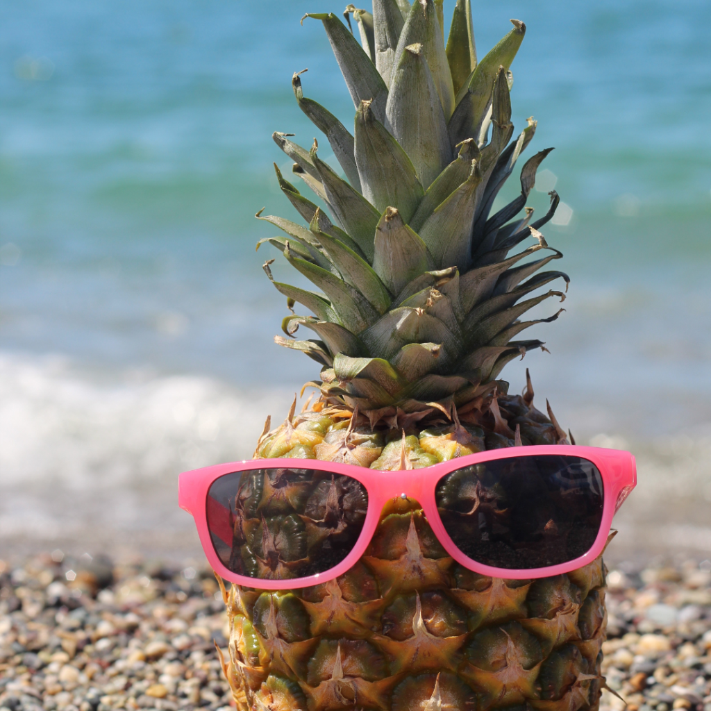 Pineapple wearing pink sunglasses on a pebble beach near the ocean
