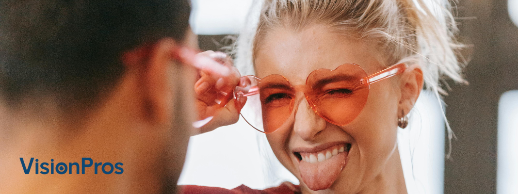 Playful woman wearing red heart-shaped sunglasses, holding the frames and sticking out her tongue, with a blurred person in the foreground and the VisionPros logo in the corner.